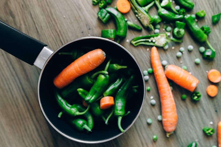 Top view of fresh carrots, chilies, and peas in and around a pot on a wooden surface.