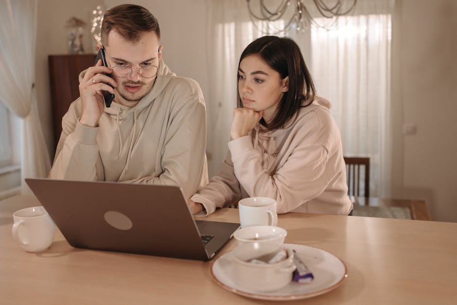 A couple in hoodies focused on work using a laptop and smartphone at home.