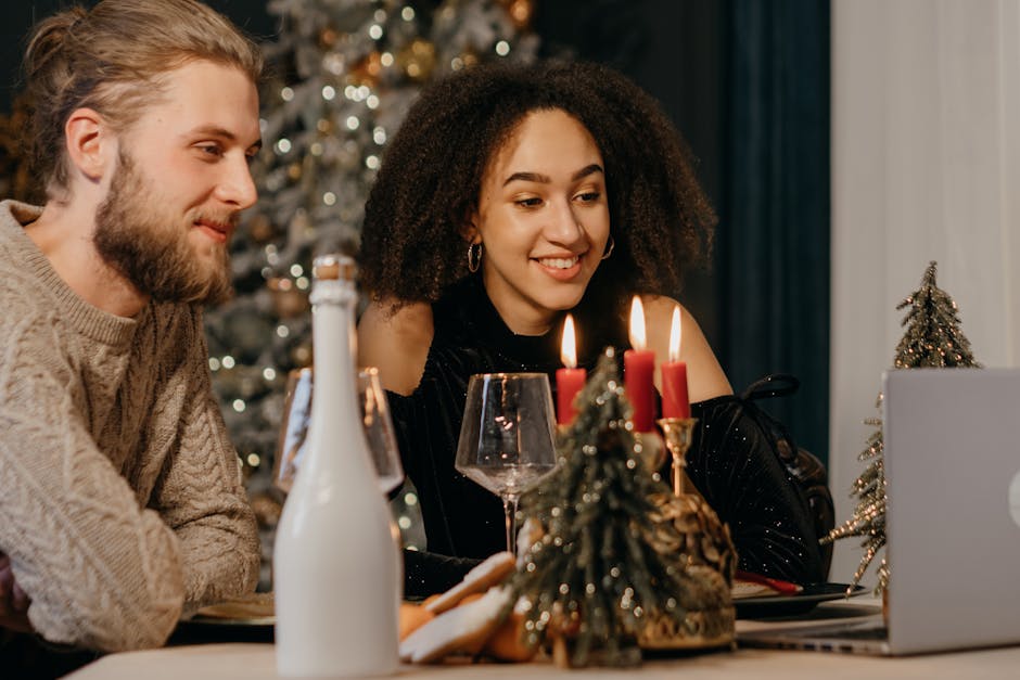 An interracial couple enjoying a festive holiday dinner with candles, wine, and a laptop for a virtual celebration.