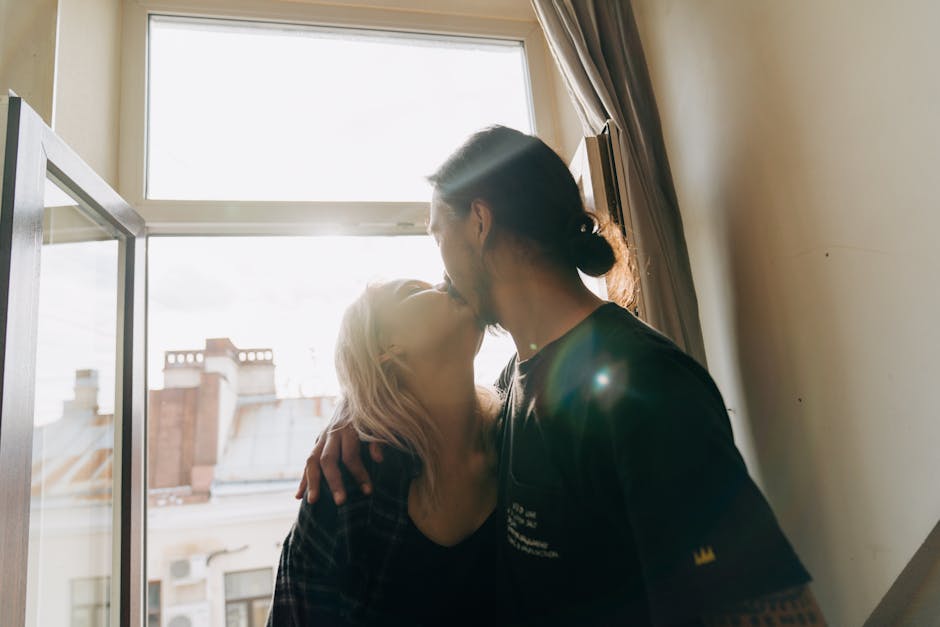 A couple sharing a romantic kiss indoors with sunlight streaming through a window.