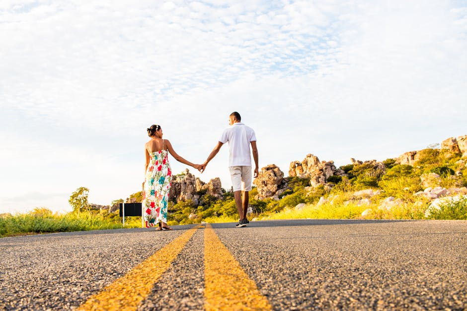 A couple holds hands while walking on a sunny countryside road, surrounded by nature.