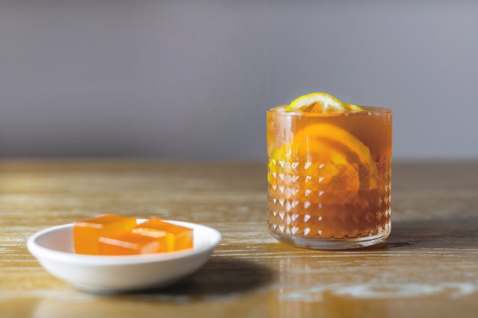 A glass of iced black tea with lemon slices and orange jelly cubes on a wooden table.
