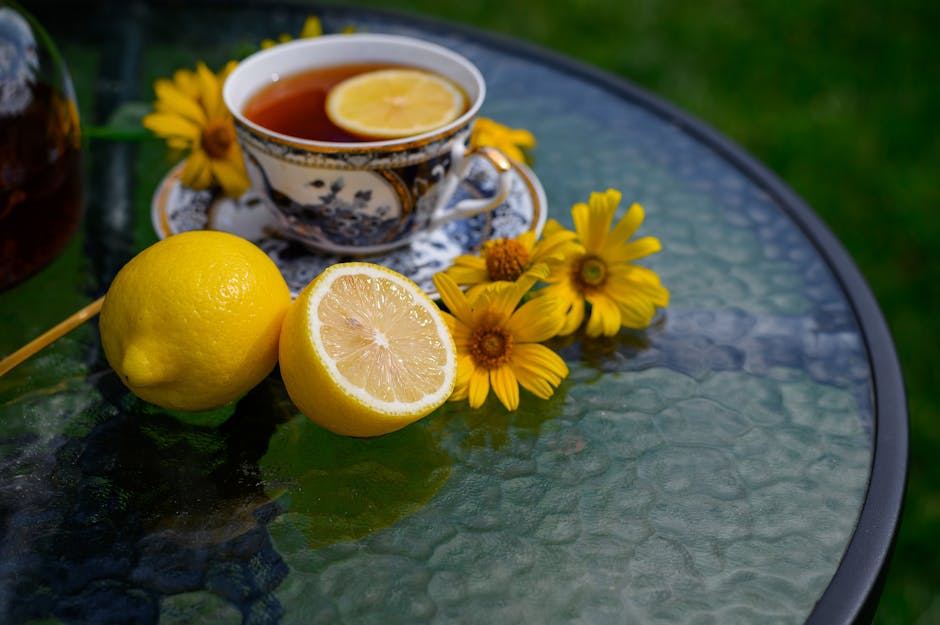 A refreshing cup of lemon tea on a glass table surrounded by yellow flowers and sliced lemon.