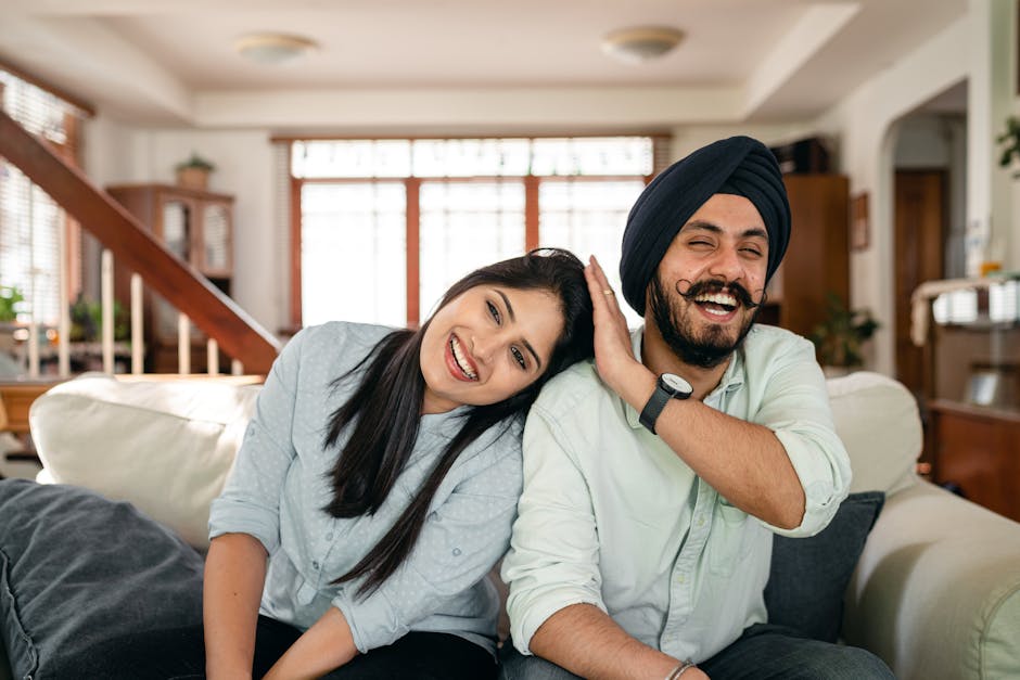 Young happy casual Indian woman leaning on shoulder of boyfriend in traditional turban while having fun together in cozy living room