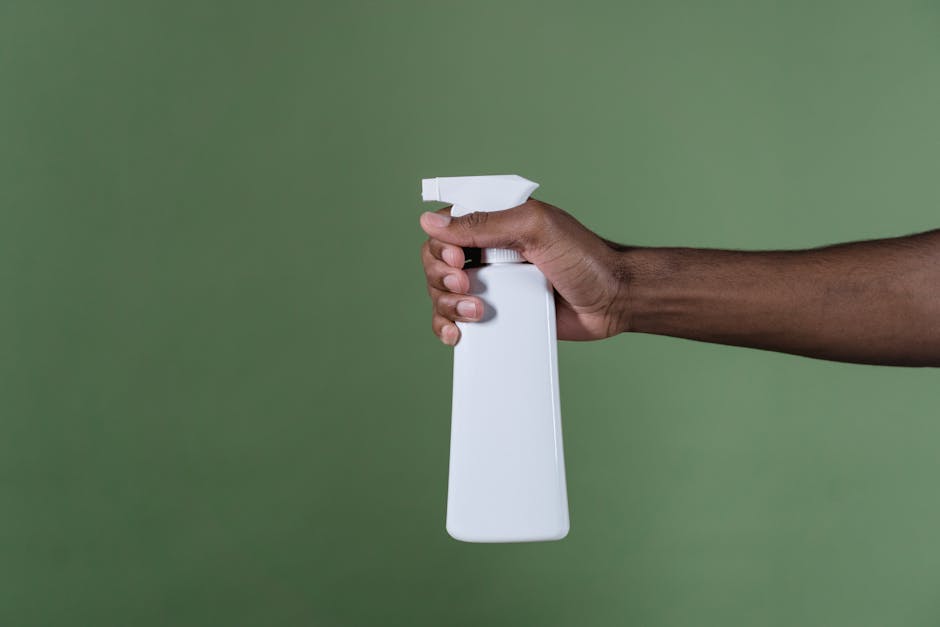 A hand holds a white spray bottle against a green background, perfect for cleaning product concepts.