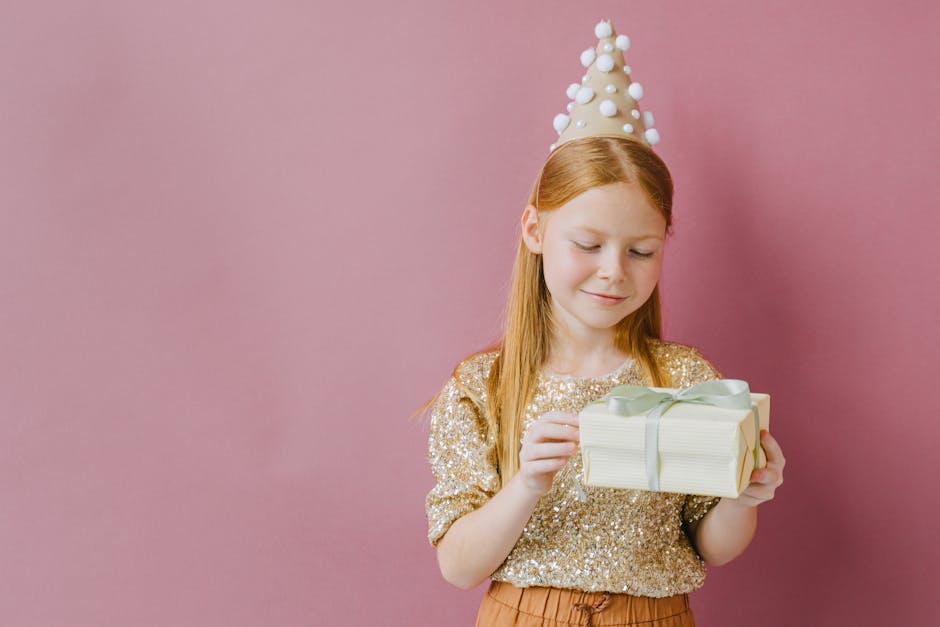 Young girl in glittery top and party hat holds a gift, set against a pink backdrop, radiating joy.